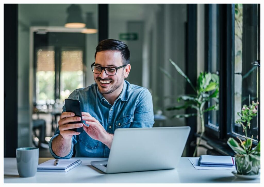 A man on his laptop looking happily at his phone.